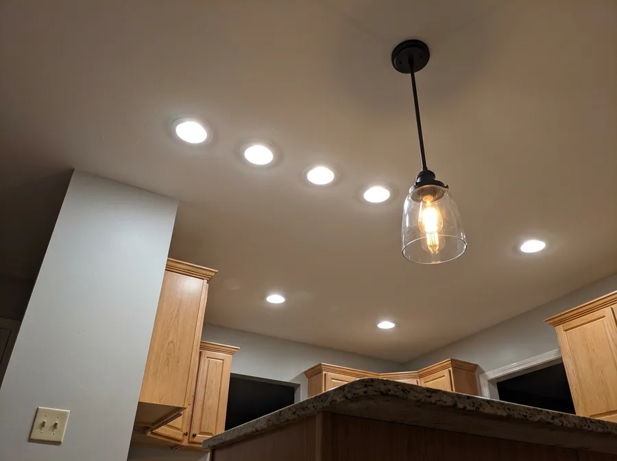 A row of recessed ceiling lights and one pendant fixture installed, wired and switched on in a finished suburban kitchen, seen from below at a slight upward angle, warm even light and no ladder clutter